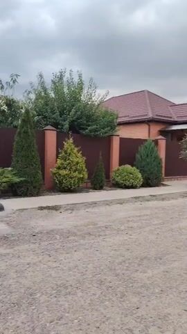 The video shows a stationary view of a house with a red roof, a fence made of brick and dark brown panels, and various green shrubs and trees. The house is a single-story structure with a covered entrance. A gravel road is in the foreground. The sky is overcast and cloudy. The camera is positioned to the side of the road, looking towards the house and its yard.