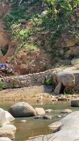 A person rides a motorcycle over a rocky path next to a river. The motorcycle is red and has luggage strapped to the back. The path is made of rocks and dirt, and the river is full of large boulders. There are trees and vegetation on the hillside in the background. The sun is shining, creating shadows.