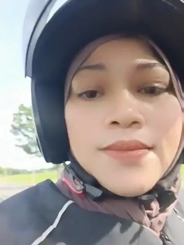 A woman wearing a hijab and a black helmet is on a motorcycle. She is wearing a dark jacket with white stripes. The background shows a field of green grass and trees. The sky is blue and cloudy. The video is a selfie from the woman's perspective.