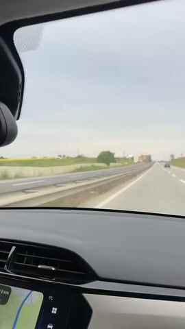 The video shows a driver's point of view while traveling on a highway. The sky is overcast and the view includes guardrails, grassy areas, and various roadside structures like billboards, buildings, and utility poles. There are other cars on the highway, including a dark-colored car and a white van in the distance. Inside the car, the dashboard is visible, featuring a navigation screen displaying a map. Music is audible throughout the video.