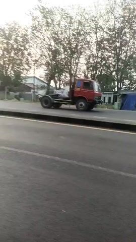 The video shows a road scene with a red truck driving on the right side of the road. There are trees and buildings in the background. A traffic light is visible on a pole. Several motorcycles are also seen on the road, some with riders. In one frame, two people on a motorcycle are wearing blue shirts. Another motorcycle is visible in the foreground, moving. There are also some flags on poles in the background. The overall environment appears to be an urban or semi-urban setting, possibly in Indonesia given the flag colors. The lighting suggests it might be daytime, possibly morning or late afternoon.