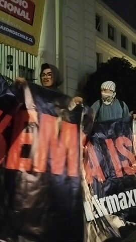 The image captures an outdoor night scene in Ñuñoa, Chile, depicting what appears to be a public demonstration or display. Three individuals are visible, with two prominently holding a large black banner.

The person on the left is wearing a light-colored keffiyeh (headscarf) and a light blue top. The person in the center wears a dark hooded sweatshirt and glasses, looking directly towards the camera. The head of a third individual is partially visible behind the banner to the right.

The main object is a large black banner made of a flexible material, possibly plastic or vinyl, featuring bold red and white text. The red text clearly shows "MAX," and white text below it, partially visible due to folds, reads "MAXPALESTINA.CL."

In the lower-left corner, a partially obscured cardboard box is visible with text including "NACIONAL" and "RAZONESDECHILE.CL." The background features a multi-story building with illuminated windows, characteristic of an urban environment at night. The scene is illuminated by artificial light sources, indicating it is dark outside. The overall impression is one of individuals engaged in an organized public message display.