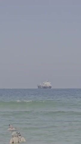 A long shot captures a wide expanse of ocean under a hazy sky. In the foreground, several people are visible in the water, some appearing to be swimming or wading. Further out, a large cargo ship is sailing across the horizon. The waves are gentle, and the overall atmosphere suggests a calm day at the beach.