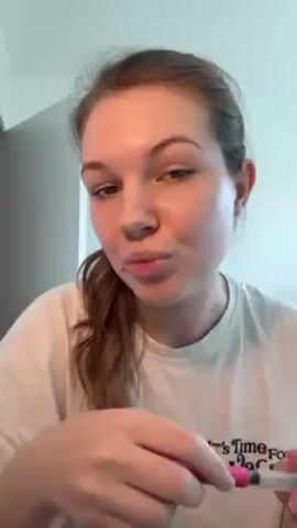 A woman in a white t-shirt is standing indoors and applying makeup to her eyebrow. The t-shirt has the phrase "It's Time For A Vacation" printed on it. The woman has green eyes and her brown hair is pulled back into a ponytail. She is holding a makeup brush with a pink handle. The background consists of a white wall and a darker grey wall. Music is playing in the background.