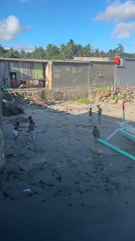 A vertical, eye-level shot captures a sunny beach scene with children playing in the sand. In the foreground, the dark, wet sand shows footprints and debris, transitioning to a lighter, drier sand area where four children are engaged in activity. Two children are on the left, one appearing to squat and the other standing. Two more children are on the right, one on his knees and another standing with a stick. In the background, a simple, weathered building with a green section and a concrete section stands. A ladder is visible against the building. A boat with a green and white hull is partially visible on the right side of the frame. Lush green foliage and palm trees are in the distance under a bright blue sky with scattered white clouds.