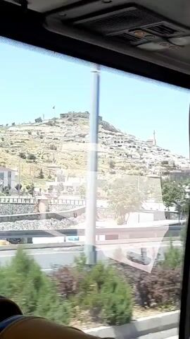The video shows a view from inside a vehicle, looking out a window at a sunlit, hilly landscape. The foreground shows the interior of the vehicle, including part of a person's arm and hand on the right side, and the top of the vehicle's interior with vents. The middle ground features a paved road with multiple lanes, some green vegetation, and a round grassy island with some trees and bushes. In the background, a large hill is covered with a cluster of buildings that appear to be ancient or historic, with some structures resembling houses and a prominent building with a minaret, possibly a mosque. At the very top of the hill, there are ruins of a fortress or castle with a flag flying. The sky is clear blue. The scene suggests a journey through a historically significant area.
