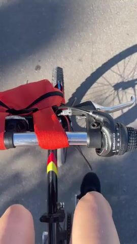 The video shows a first-person view of someone riding a bicycle on an asphalt surface. The handlebars are visible, with a red bag attached. The bicycle has black and yellow accents. The rider's legs and feet wearing black sneakers are visible. The shadow of the bicycle and rider is cast on the ground due to the bright sunlight. The bicycle has Shimano gears.