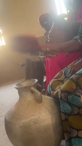 In an indoor setting, two women are seated.  The woman closest to the camera wears a teal, yellow, and brown patterned dress, and her hands are visible. The other woman, seated behind, wears a patterned shirt, a pink skirt, and a headscarf. In front of the first woman is a large, gray, clay pot with a handle.  The woman uses a red and black woven object to strike the pot rhythmically, creating a musical sound. Behind the second woman, a drum-like object rests on a wooden stool. The scene is brightly lit by sunlight streaming in from an unseen window.