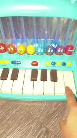A close-up shot shows a child's hand pressing keys on a toy piano. The piano is turquoise and shaped like a hippopotamus. The keys are white and black. Above the keys are several colorful, round characters inside clear tubes, which light up as the keys are pressed. Some of the characters are red, orange, yellow, green, light blue, dark blue, purple, and pink. The keys have some pink marks on them, possibly from marker. The piano is resting on a wooden floor.