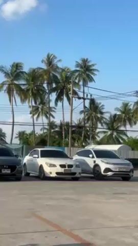 The video shows four cars parked in a lot with palm trees in the background. The cars are a dark gray Mazda, a white BMW sedan, a white crossover SUV, and another light-colored car partially visible to the right. The sky is blue with some clouds. There are utility poles and power lines near the palm trees. The setting appears to be a sunny, tropical location.