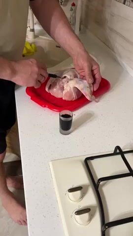 A man is shown close up, standing in a kitchen, injecting a dark liquid from a small shot glass into a chicken. He is using an injector with a black handle and a needle. The chicken is on a red plastic tray on a white speckled countertop next to a stove. The man has a wedding band on the ring finger of his left hand. The shot glass appears to be frosted. He is wearing a beige t-shirt and black shorts. The kitchen has white tiling and a sink visible in the background.