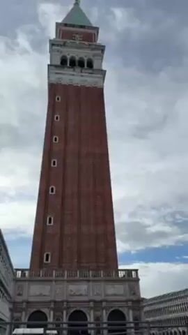 The video shows a low-angle view of the bell tower (Campanile) of St. Mark's Basilica in Venice, Italy.  The camera pans right to show the Basilica itself, partially covered in scaffolding and mesh. The sky is cloudy with patches of blue visible.  There are no people visible in the video.