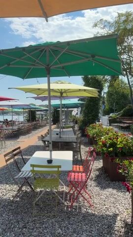 The video shows an outdoor seating area of a restaurant or cafe with a waterfront view. There are multiple tables and chairs set up on a gravel surface, many of which are shaded by large, colorful umbrellas in various shades of green, yellow, orange, pink, and white. A wooden walkway extends from the seating area towards the water, with more tables and chairs visible beyond. Several people are seated at the tables, some interacting, others appearing to be dining or relaxing. In the background, the water stretches out to a distant shoreline with trees and hills. The sky is blue with some scattered clouds. Some people are visible near the water's edge, with one person in the water and another on a small wooden pier. There are also some lounge chairs and other beach-like amenities near the water. The overall scene depicts a pleasant outdoor dining and relaxation spot on a sunny day.