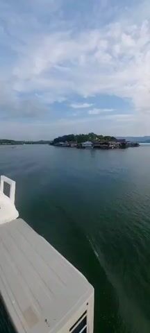 The video shows a view from a boat on a body of water, likely a river or lake. In the distance, a small island settlement with stilted houses and buildings is visible. The houses are clustered together and appear to be on the water's edge or directly over it. The water is calm, with small ripples suggesting the boat is moving. The sky is overcast with scattered clouds. As the video progresses, the camera pans to the left, revealing more of the shoreline with similar stilted houses and lush green vegetation on the hills behind them. The overall impression is of a quiet, waterfront community.