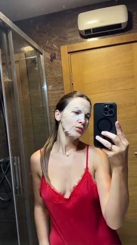 In a bathroom with marble-patterned walls and a wooden door, a woman with her hair in a ponytail is taking a selfie in the mirror. She is wearing a red slip with lace trim and a thin necklace. On her face, she has a sheet mask, and she is in the process of removing it from her chin. She holds the mask in her right hand, pulling it upwards and away from her face, while her left hand is near her chin. Her expression appears neutral, perhaps a bit focused on the task. A smartphone is held in her right hand, capturing the image. In the background, a glass shower door with a showerhead is visible to the left, and a white heating unit is mounted on the wall above the door. The lighting suggests it is daytime.