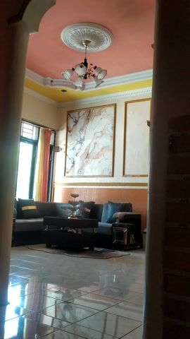 This media file showcases an interior view of a living room in a home located in Desa Penggalang, Kecamatan Adipala, Indonesia. The scene is brightly lit, suggesting it is daytime. The room is furnished with a modern, dark gray sectional sofa and a matching rectangular coffee table. A decorative tiered stand is placed on the coffee table. The walls are adorned with large framed artwork, one featuring a marble-like pattern with gold accents, and another with a simpler, textured design also framed in gold. The ceiling is a prominent feature, painted a soft pink with a yellow border and decorated with an ornate white ceiling rose from which a multi-armed chandelier hangs. The floor is made of glossy, reflective tiles that mirror the room's elements. Sunlight streams in from a window on the left, casting light and shadows and illuminating the curtained window. The overall ambiance of the room appears to be one of comfort and modern elegance. There is no visible text in the media file.