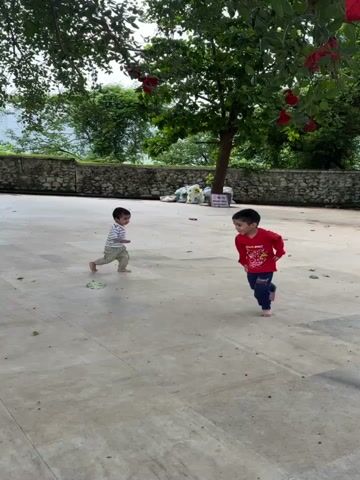 Two young boys are running on a paved outdoor area. The boy in the foreground is wearing a red shirt and blue pants. The boy in the background is wearing a striped shirt and tan pants. There is a large tree with red decorations in the background, along with a stone wall and some foliage. The sky appears to be overcast.