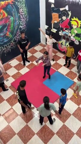 In an indoor space with a checkered floor, several children and young adults are engaged in a dance activity. A young girl in a floral shirt and dark pants is the central figure, performing a dynamic dance move with her arms outstretched. She is positioned on a red mat, which is part of a larger puzzle-like mat arrangement on the floor, with a blue mat adjacent to it. Other participants, dressed in casual attire, are standing in a circle around the central mat, observing and possibly waiting for their turn. In the background, the walls are decorated with vibrant murals, including an abstract depiction of a dancer on the left and characters from the anime series Naruto on the right. A sound system, including speakers, is visible on the left side of the frame. The overall scene suggests a dance class or practice session taking place in a creatively decorated studio.