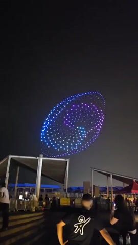 The video shows a night sky with a large, oval-shaped drone display. The display is made up of many small, colored lights that form a swirling pattern. The pattern is primarily purple and blue, with some pink and white lights mixed in. The drones are arranged in a grid-like formation, and their lights are synchronized to create the illusion of movement. In the foreground, there are several people watching the display. They are silhouetted against the bright lights of the drone show, and their faces are not visible. The environment appears to be an outdoor gathering space, possibly a park or a waterfront, with some structures visible in the background. The overall impression is one of a festive and impressive aerial performance.
