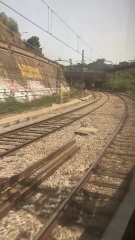A high-angle, first-person perspective shot from a moving train shows a double set of train tracks curving into the distance. The tracks are flanked by gravel and sparse vegetation. To the left, a concrete retaining wall is covered in graffiti. Overhead, a network of electrical wires and support poles stretch across the scene. In the distance, bridges and overpasses can be seen, along with more vegetation. A speed limit sign indicating "30" is visible near the tracks. The overall environment appears to be urban or suburban. The train is moving forward, indicated by the blurred motion of the tracks and the perspective shift.