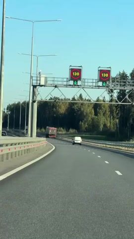 The video shows a view from a vehicle driving on a highway. Two digital speed limit signs are visible overhead, each displaying "130" and a downward green arrow. The highway has multiple lanes and is bordered by a guardrail and trees. A red truck and a white car are ahead on the road. The sky is clear and blue.