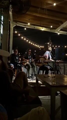Under a string-lit patio, three musicians perform for a small crowd. The band consists of a drummer, an electric guitarist, and an acoustic guitarist who also sings into a microphone. Patrons lounge in the dark foreground, the focus being on the performance. A glass of liquid sits on a nearby wooden table, part of the patio's casual setup. The atmosphere is intimate and dimly lit, with a ceiling fan above adding to the relaxed vibe.