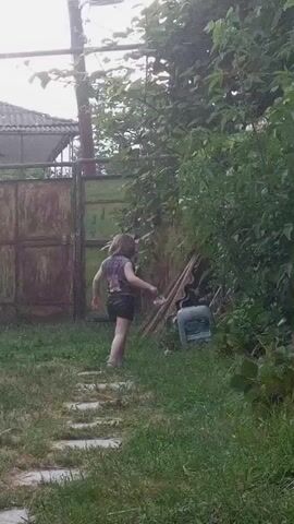 A young child is walking on a path of stepping stones in a backyard. The child is wearing a purple shirt with a graphic design and dark shorts. To the right of the child, there is a large bush and some discarded items, including what appears to be a blue and gray appliance. In the background, there is a rusty metal gate and a fence. The ground is covered in grass.