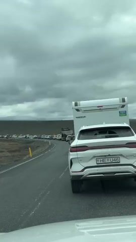 The video, filmed from inside a vehicle, captures a long line of traffic on a road through a barren landscape under an overcast sky.

At the beginning, a white SUV with a license plate partially visible as "HE U" is directly ahead, followed by a white camper van. A long queue of vehicles stretches into the distance along the road to the left. The camera then pans left, showing more of the extensive line of cars and the desolate, dark earth with sparse vegetation.

As the camera continues to pan, a distinctive modern white bridge with two tall, rectangular pylons becomes visible in the distance. Beyond the bridge, snow-capped mountains can be seen under the cloudy sky. The line of cars appears to be leading towards and across this bridge. The overall scene suggests a significant traffic delay or queue in an open, remote area of Iceland.