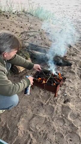 A person kneels on a sandy beach, tending to a small barbecue grill filled with burning wood and pinecones. They are using a portable butane torch to ignite the fire. Smoke billows from the grill, and the person is wearing a green hooded jacket and blue jeans. In the background, a large log and some sparse vegetation are visible. The overall scene depicts an outdoor activity of starting a campfire.