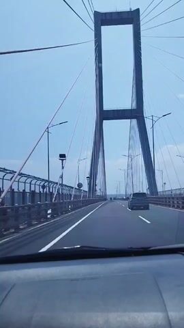 The video shows a first-person view from a car driving across a cable-stayed bridge. The sky is clear and blue. The bridge has tall, gray towers with many cables extending to the roadway. There are other cars on the bridge. On the left side of the road, there is a railing and street lights. A speed limit sign is visible.