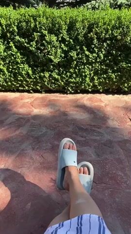 The video is a first-person perspective shot focusing on a person's feet resting on a stone patio. The person is wearing light blue slides and striped shorts. Behind the feet is a neatly trimmed green hedge, and beyond that, tall trees and the upper portion of a building are visible. The scene appears to be outdoors on a sunny day.