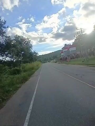 A first-person view from a motorcycle riding down a paved road. The road has a white dashed line on the left side. On the right side of the road is a building with red and white accents and several trees in front of it. On the left side of the road is a grassy area and trees. The sky is blue with white clouds and the sun is shining. Two people are riding on the motorcycle, both wearing helmets and dark clothing. They are moving away from the camera.