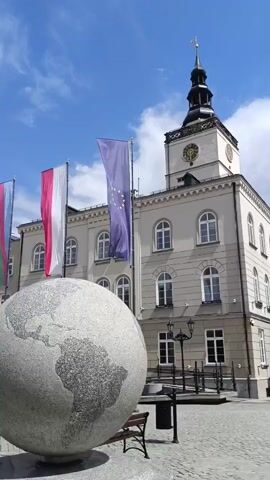 The video shows a public square with a large, spherical sculpture in the foreground that resembles a globe, etched with continents. Behind the sculpture, several flags are visible, including the Polish flag and the European Union flag. A building with a clock tower stands in the background. The scene is set outdoors on a sunny day with a blue sky and some clouds.