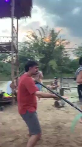 The video shows a man performing a poi dance on a sandy riverbank. He's wearing a red shirt and grey shorts, twirling a long stick with glowing green and grey blades on either end. Several other people are present. Some are seated on makeshift chairs under a bamboo structure, while others stand nearby. The background reveals a river flowing gently and lush vegetation, with the sky displaying the colors of a setting sun. A metal frame spelling "ZOO" is visible further behind.