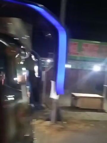 The video shows the exterior of a dark-colored vehicle, possibly a bus, at night. The vehicle has graphics on its side, including a silhouette of a person wearing a hat and text that is partially visible. There are lights on the vehicle and in the background, suggesting it is in an urban or commercial area. The camera appears to be moving, as indicated by the slight blur in the footage. The overall impression is that of a moving vehicle at night, possibly captured by someone inside or alongside it.