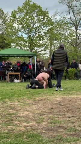 In an outdoor setting on a grassy field, two people are engaged in wrestling on the ground. One person is bare-chested with dark pants and is on top, while the other is wearing dark clothing. A crowd of people of varied ages surrounds them, watching the wrestling match. A green tent is set up nearby, under which some individuals are seated at a table. Trees provide a backdrop to the scene, and the sky is overcast. A man in a dark vest walks past the camera, observing the wrestling match.