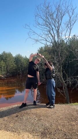 Two individuals, a male and a female, are positioned on a pebbled edge overlooking a body of water with a distinct red hue. A leafless tree stands to their right, with evergreen trees forming a backdrop in the distance against a clear blue sky. The male, dressed in a black t-shirt and black shorts, has his left arm raised and arched above his head. The female, sporting a black vest over a patterned shirt and blue jeans, mirrors the gesture with her right arm, their hands meeting to form a heart shape.