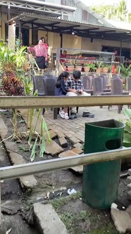 The video shows an outdoor scene with people and a building. In the foreground, a metal railing partially obstructs the view. Beyond the railing, there is a paved area with broken concrete slabs and some greenery. To the right, a green trash receptacle is visible. In the middle ground, two young people, a male and a female, are seated on plastic chairs, facing each other and looking at a mobile phone. The male is wearing a plaid shirt, and the female is wearing a dark blue jumpsuit. Behind them, there are rows of empty plastic chairs. The background features a building with a corrugated metal roof and a porch area. Several adults are present on the porch. One woman in a red and white striped shirt is gesturing with her hands as she walks down stairs. Other individuals are seated or standing on the porch. The environment appears to be a casual outdoor gathering space with a garden area and seating arrangements.
