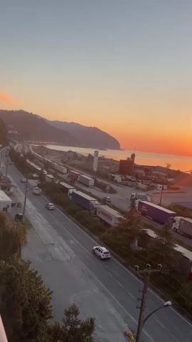 The video displays a high-angle view of a coastal road at sunset. A highway runs parallel to a coastline, with a line of parked trucks on the side of the road. The sun is setting over the ocean, casting a warm orange glow across the sky and water. Mountains are visible in the distance. Several cars are driving on the highway. The overall scene is peaceful and scenic, with a sense of quietude as the day draws to a close.