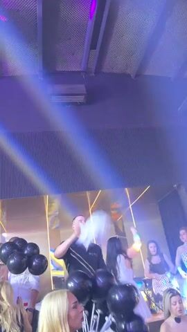 A club atmosphere is captured, with people and objects visible under multicolored light beams. The scene includes a man holding a bouquet of black balloons, surrounded by women. Paper is thrown into the air. Neon lights beam across the scene, and a large screen displays figures.
