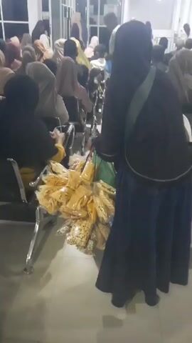 A woman wearing a black hijab and a dark blue skirt stands in a waiting area, facing away from the camera. She is holding a bunch of bagged snacks. She appears to be offering them to people seated in chairs in front of her. The waiting area is filled with many people, most of whom are wearing hijabs. The chairs are metal and arranged in rows. There is a sign that says "POLI MATA" which likely indicates an eye clinic. The lighting is artificial and somewhat dim. The floor is tiled.