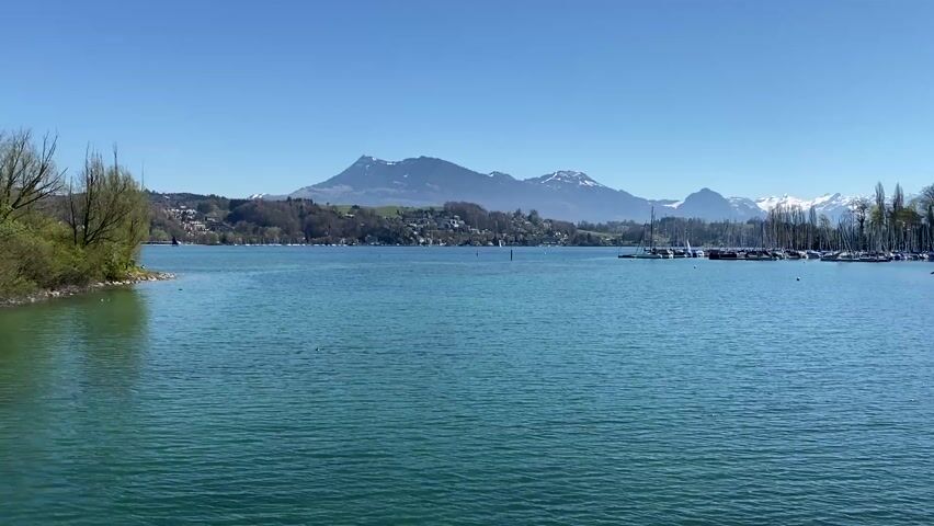The video shows a wide shot of a lake with a clear blue sky overhead. The water is a turquoise color with gentle ripples on its surface. On the left, a small, green, tree-lined shoreline extends into the lake. In the distance, a mountain range is visible with some snow-capped peaks.  On the right side of the frame is a marina filled with sailboats, their masts creating a dense vertical pattern against the landscape. A small village is nestled on the hills along the lake's edge.