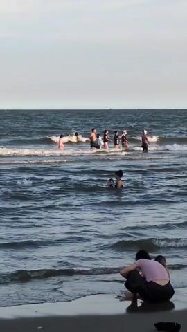 The video shows a beach scene with several people in and near the water. In the foreground, a person is crouched on the sand, facing the water. Further out in the ocean, a group of people are standing in the waves, some appearing to be children. Closer to the shore, a person is in the water holding what appears to be a child. In the distance, a lone person is visible further out in the water. The ocean is choppy with visible waves. The sky is overcast.
