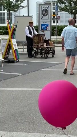 A man in a hat plays a street organ, which is on a cart with wheels. He is wearing a white shirt, a black vest, and black pants. To his left is a stand with oversized colored pencils. To his right are two road signs, one with a "ZONE" label and a person icon, and another with a "ZONE" label and a car and child icon. A man wearing a light blue shirt and shorts walks from right to left in the background. In the foreground, a large pink balloon bobs in the lower right corner. The street is paved, and there are white buildings and trees in the background.