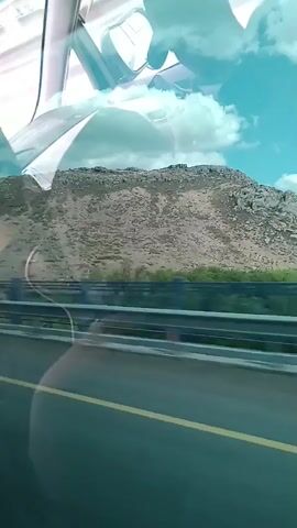 The video shows a view from inside a vehicle, likely a car or bus, looking out the side window. The perspective is slightly angled, indicating movement. In the foreground, a yellow line is visible on a road, suggesting a highway or main road. A metal guardrail runs parallel to the road. Beyond the guardrail, there are some green trees and shrubs. Further in the background, a large, rocky, and barren mountain slopes upwards against a bright blue sky with fluffy white clouds. The interior of the vehicle is reflected in the window, showing parts of the car's interior, including a side mirror and the ceiling. The overall impression is a scenic drive through a mountainous landscape.