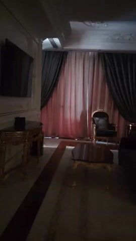 The video pans across a dimly lit bedroom. The room appears to be luxuriously decorated, with ornate trim on the walls and ceiling. In the foreground, a plush rug covers the wooden floor. A dark, ornate coffee table sits in the center of the room. To the right, a dark sofa is partially visible. Behind the coffee table, there's a single armchair with dark upholstery and gold trim, facing a large window covered by heavy, layered curtains of pink and dark gray. Further back, a large bed with a white headboard and white bedding is the dominant feature. Beside the bed, a dark wooden nightstand holds a lamp with a white shade. The camera continues to pan, revealing more of the bed and the detailed wall paneling, which is a creamy white with gold accents. Another dark wooden nightstand with a similar lamp is visible on the other side of the bed. A floor lamp is also present. The video ends by panning back towards the bed and the curtains.