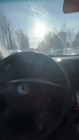 The video shows the driver's perspective inside a Skoda car, driving on a snow-lined road on a bright, sunny day. Other cars are visible on the road ahead, with leafless trees lining the sides. The Skoda's steering wheel, dashboard, and infotainment screen are visible in the foreground. The windshield is partly obscured with streaks.