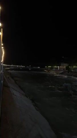 The video shows a dark, outdoor scene at night. A river flows towards the viewer's right, with visible ripples and some white water suggesting movement. The riverbanks are lined with concrete or stone walls. On the right bank, a series of string lights are visible, illuminating a pathway and some trees. A building or structure with lights is also visible in the background on the right. The sky is completely black, indicating it is nighttime. The left bank is less illuminated, showing a concrete slope leading down to the water. There are no people clearly visible in the frame. The overall atmosphere is dark and somewhat tranquil, with the gentle glow of lights contrasting with the darkness.