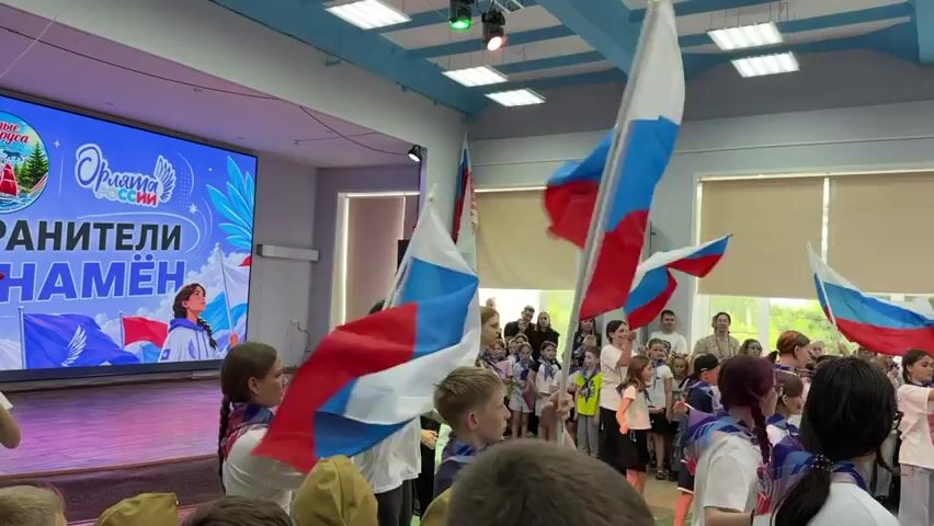 A group of children, some waving Russian flags, are gathered in an indoor space. A large screen displays text and images, including what appears to be a patriotic message and a young person. The children, some wearing scarves, are facing the screen, and others are in the foreground, obscuring some of the view. The environment appears to be a school or community hall, with lighting fixtures visible on the ceiling. The flags are a prominent feature, with their red, white, and blue colors waving in the air. Some of the children are looking towards the screen, while others are interacting with each other. The overall atmosphere suggests some kind of event or celebration.