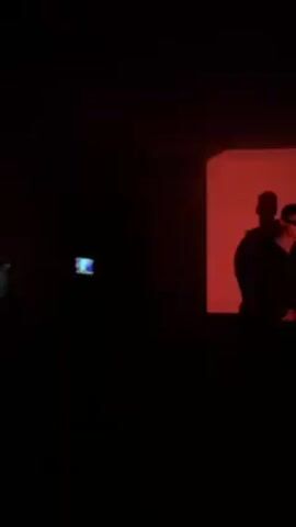 The scene is a dark room, likely indoors. A person wearing sunglasses and a dark jacket stands in front of a wall illuminated with a square of blue light. The person appears to be dancing or moving to music. The silhouette of the person is also visible on the wall. In the foreground, another individual is recording the scene with a camera or smartphone. The overall atmosphere is dimly lit and focused on the person being recorded.